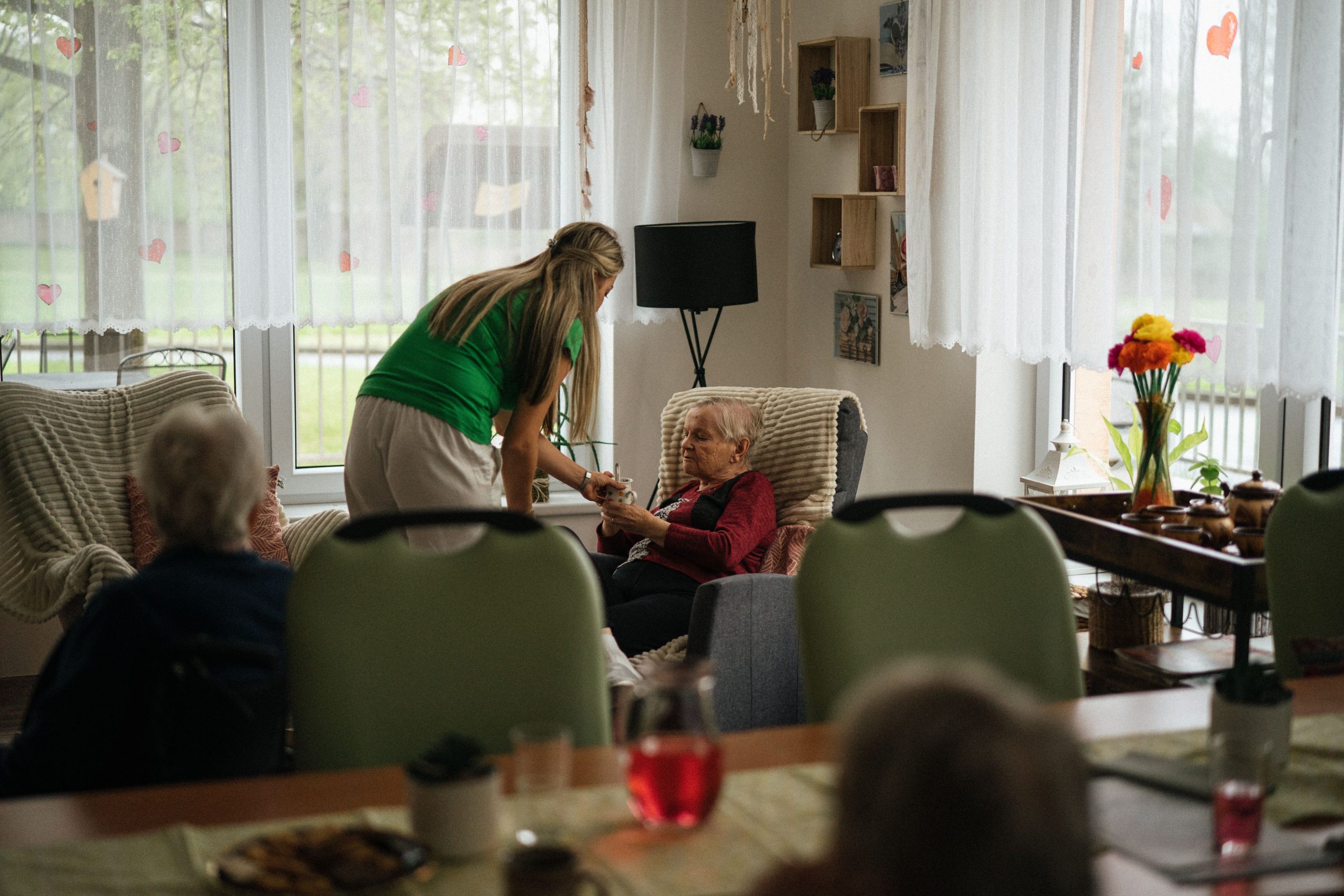 Caregiver assisting elderly women with tea in a cozy nursing home setting.